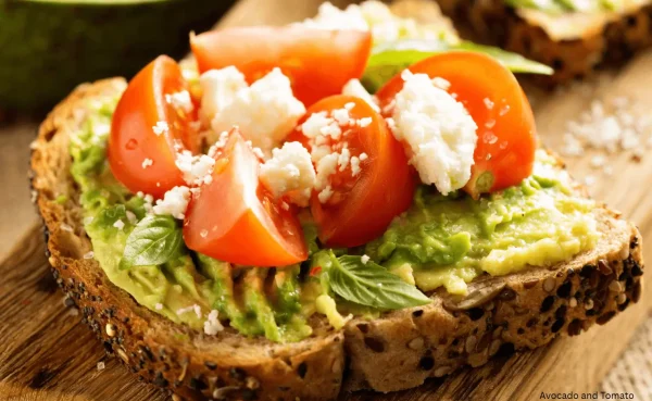 Open faced avocado sandwich topped with fresh tomatoes, greens, and crumbled cheese on whole grain bread
