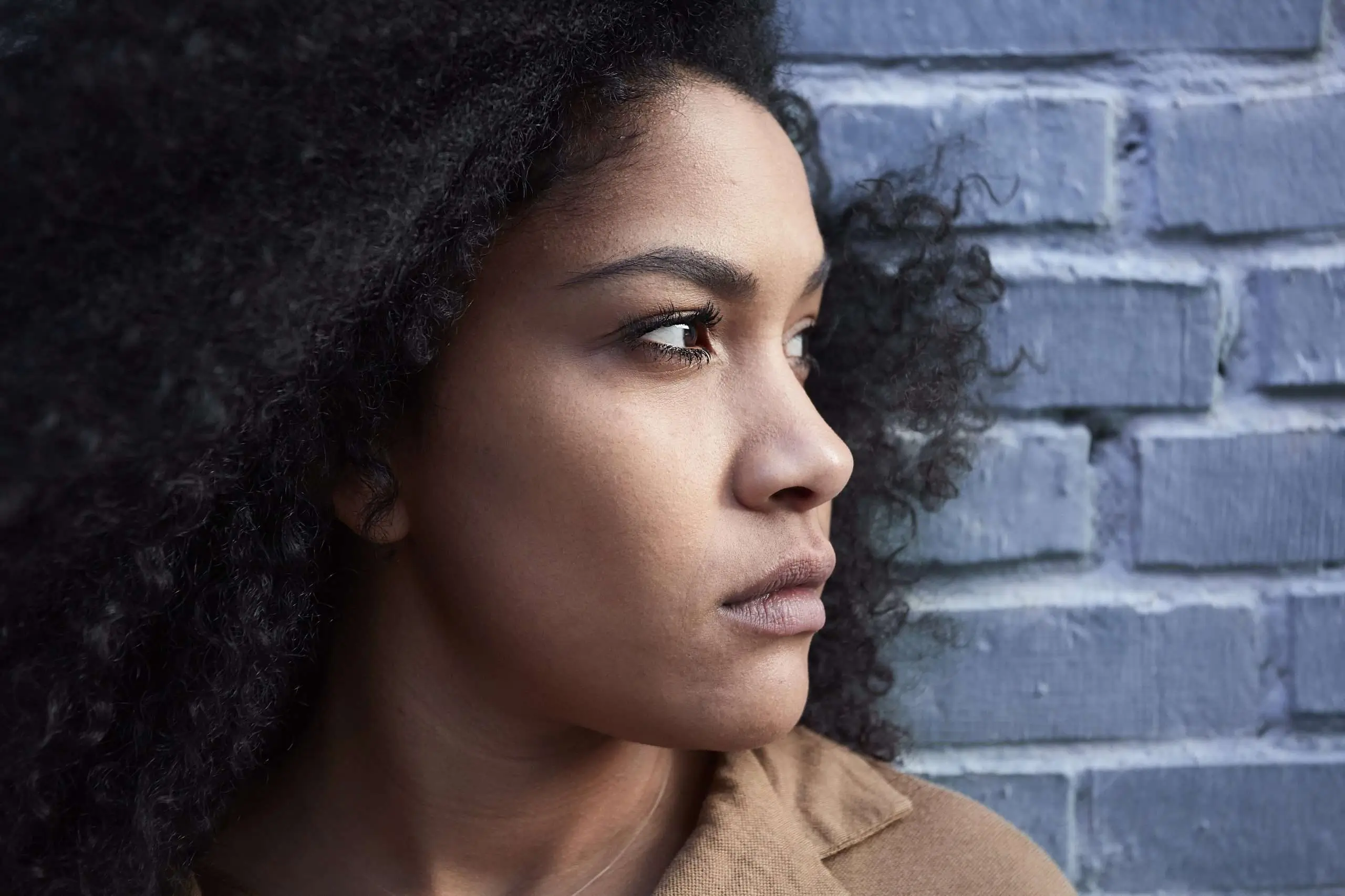 Black woman sitting against a wooden wall with a thoughtful expression for an article about the emotional side of acne.