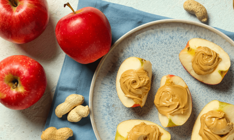 Apple slices topped with peanut butter on a plate with whole apples and peanuts nearby