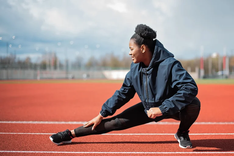 Black woman stretching on an outdoor running track for an article about how exercise can benefit skin.